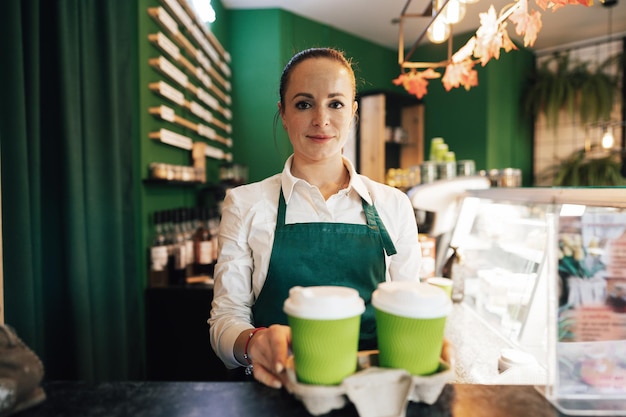 Junge Barista-Frau mit Schürze, die ein Tablett mit fertigem Kaffee hält