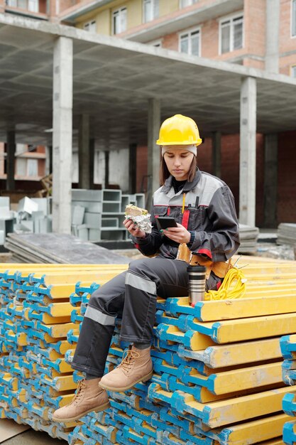 Junge Baumeisterin in Arbeitskleidung mit Sandwich und Smartphone