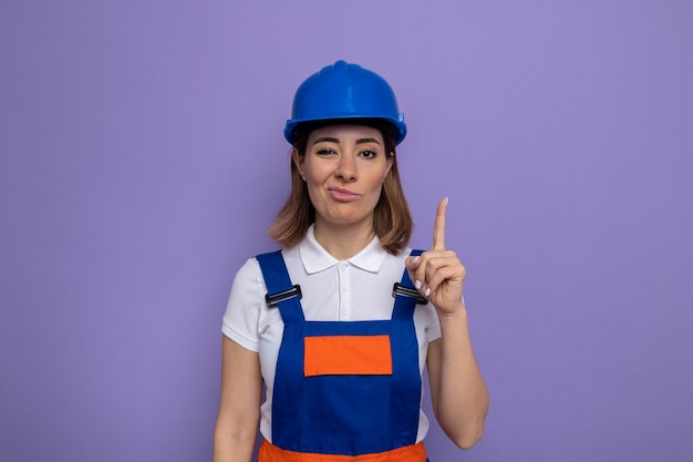 Junge Baumeisterin in Bauuniform und Schutzhelm, die einen trockenen Mund mit enttäuschtem Ausdruck macht, der den Zeigefinger zeigt, der über der lila Wand steht?