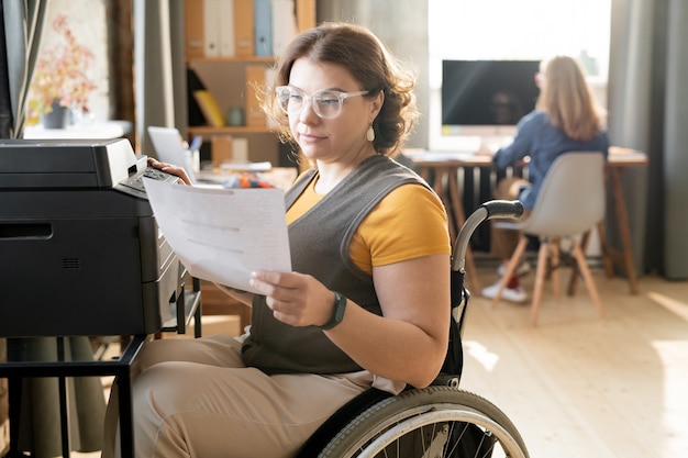 Junge behindert weibliche Büroangestellte oder Sekretärinnen, die durch Papier schauen, während sie an der Xerox-Maschine sitzen und Kopien gegen Kollegen anfertigen