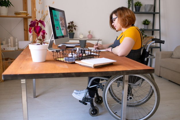 Junge behinderte Frau im Rollstuhl, der Notizen vor Computermonitor macht, während sie zu Hause während der Fernstunde am Schreibtisch sitzt