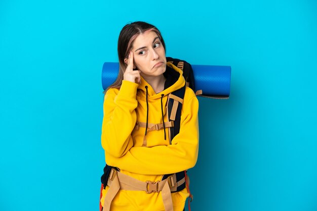 Junge Bergsteigerfrau mit einem großen Rucksack lokalisiert auf blauer Wand, die eine Idee denkt