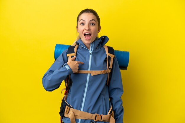 Junge Bergsteigerfrau mit einem großen Rucksack über isoliertem gelbem Hintergrund mit überraschendem Gesichtsausdruck