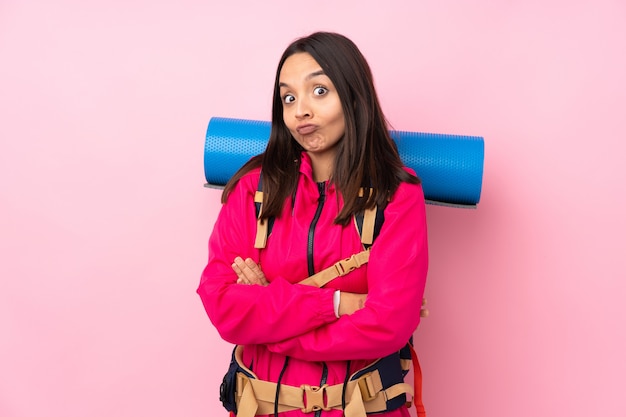 Junge Bergsteigerfrau mit einem großen Rucksack über isolierter rosa Wand, die Zweifel gestikuliert, während sie die Schultern anhebt