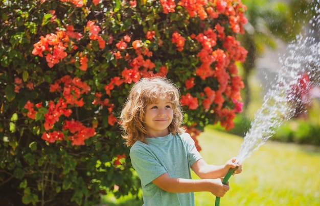 Junge Bewässerung von Pflanzen im Garten. Das Konzept der Kinderfreundlichkeit und Kindheit. Kleiner Helfer. Schönes blondes Kind, das im Sommer im Haus im Freien einen Wasserschlauch im Garten hält.