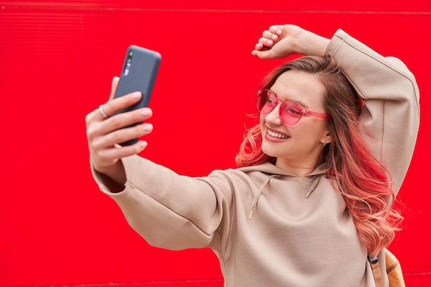 Junge Bloggerin mit rosa gefärbten Haaren, die nahe der roten Wand bleiben und Selfie durch Smartphone machen