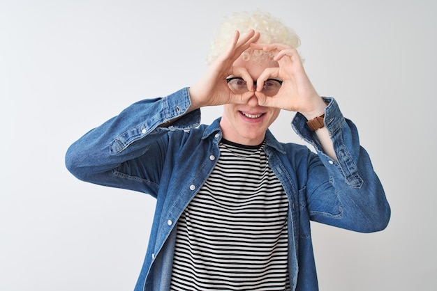 Junge blonde Albinos, der ein Jeanshemd und eine Brille über einem isolierten weißen Hintergrund trägt, macht eine ok-Geste wie ein Fernglas, steckt die Zunge aus den Augen und schaut durch die Finger.