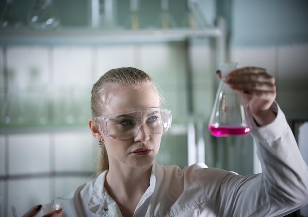 Junge blonde Frau des chemischen Labors, die eine Flasche mit rosa Flüssigkeit darin hält Frau im Fokus