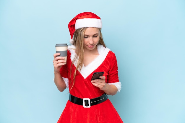 Junge blonde Frau, die als Mama Claus verkleidet ist, isoliert auf blauem Hintergrund, hält Kaffee zum Mitnehmen und ein Handy