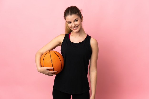 Junge blonde Frau, die Basketball auf rosa Hintergrund spielt, der mit Armen an der Hüfte aufwirft und lächelt