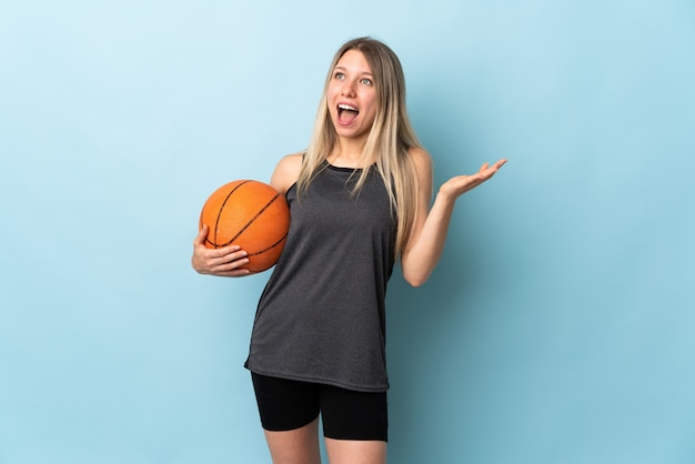 Junge blonde Frau, die Basketball lokalisiert auf blauer Wand mit überraschendem Gesichtsausdruck spielt