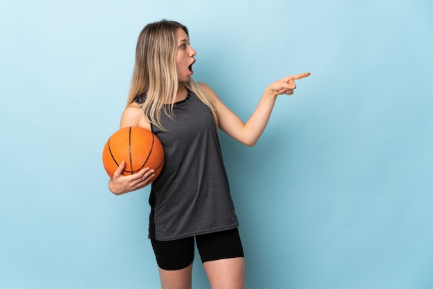 Junge blonde Frau, die Basketball lokalisiert auf blauer Wand zeigt Finger auf die Seite zeigt