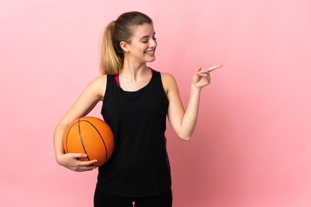 Junge blonde Frau, die Basketball lokalisiert auf rosa Hintergrund zeigt Finger zur Seite zeigt und ein Produkt präsentiert