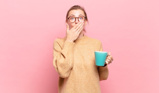 Junge blonde Frau, die den Mund mit den Händen mit einem schockierten, überraschten Ausdruck bedeckt, ein Geheimnis hält oder oops sagt. Kaffeekonzept