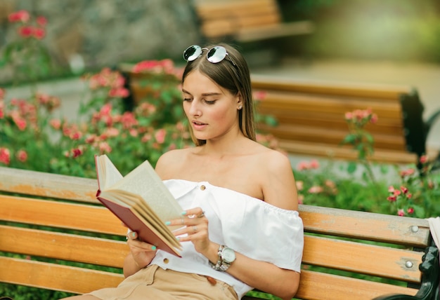 Junge blonde Frau, die ein Buch mit Begeisterung liest, während sie auf einer Bank im Park sitzt