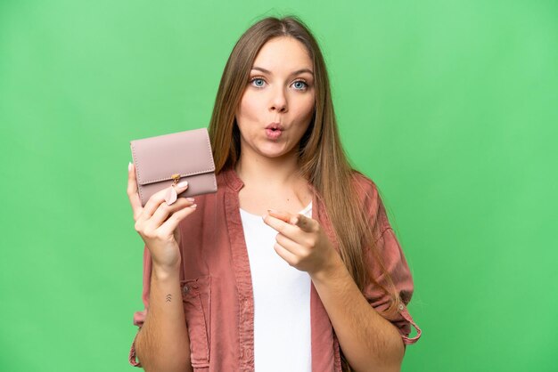 Junge blonde Frau, die eine Brieftasche über isoliertem Chroma-Key-Hintergrund hält, überrascht und nach vorne zeigend