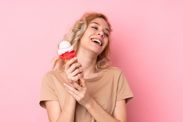 Junge blonde Frau, die eine Kornett-Eiscreme lokalisiert auf rosa Wand hält