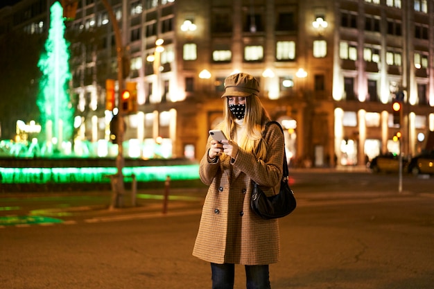 Junge blonde Frau, die eine Maske trägt, ein Smartphone benutzt und eine Nachricht schreibt, die in einer Stadt in der Nacht steht. Winteratmosphäre.