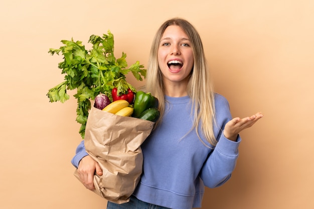 Junge blonde Frau, die eine Tasche voll des Gemüses über isolierter Wand unglücklich und mit etwas frustriert hält