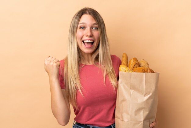 Junge blonde Frau, die eine Tasche voller Brote lokalisiert auf beige Wand hält, die einen Sieg feiert