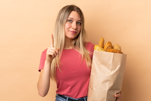 Junge blonde Frau, die eine Tüte voll Brot lokalisiert auf beige Wand hält, die mit dem Zeigefinger eine große Idee zeigt