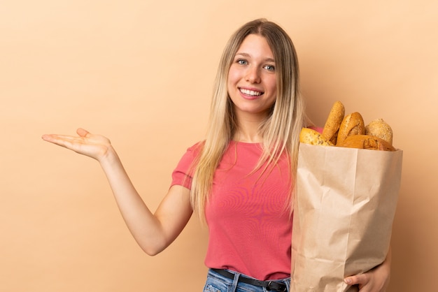 Junge blonde Frau, die eine Tüte voll Brot lokalisiert auf beiger Wand hält, die Hände zur Seite für Einladung zum Kommen ausdehnt