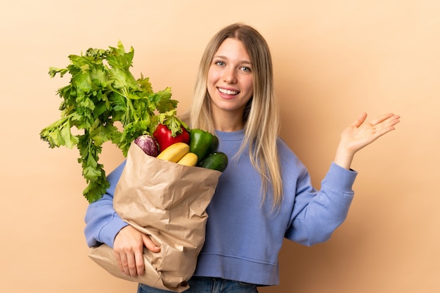 Junge blonde Frau, die eine Tüte voll Gemüse über isolierte Wand hält, die Hände zur Seite für Einladung zum Kommen ausdehnt