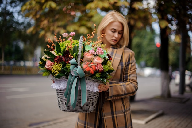 Junge blonde Frau, die einen großen Weidenkorb der Blumen gegen die Stadt hält