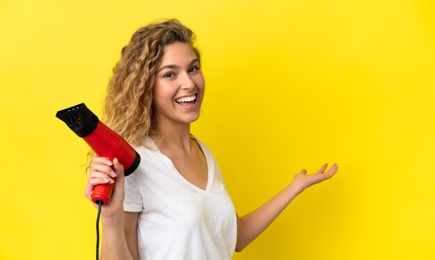 Junge blonde Frau, die einen Haartrockner isoliert auf gelbem Hintergrund hält und die Hände zur Seite ausstreckt, um zum Kommen einzuladen?