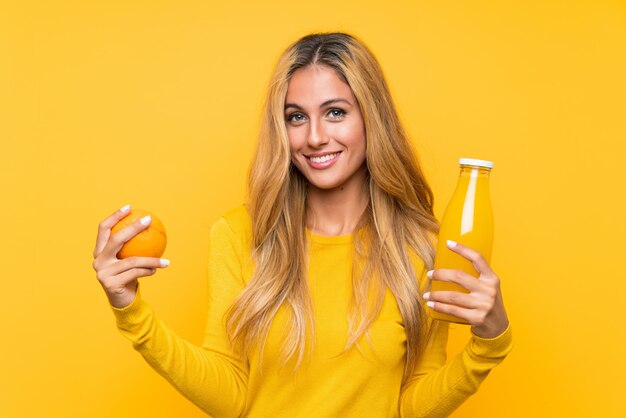 Junge blonde Frau, die einen Orangensaft über gelbem Hintergrund anhält