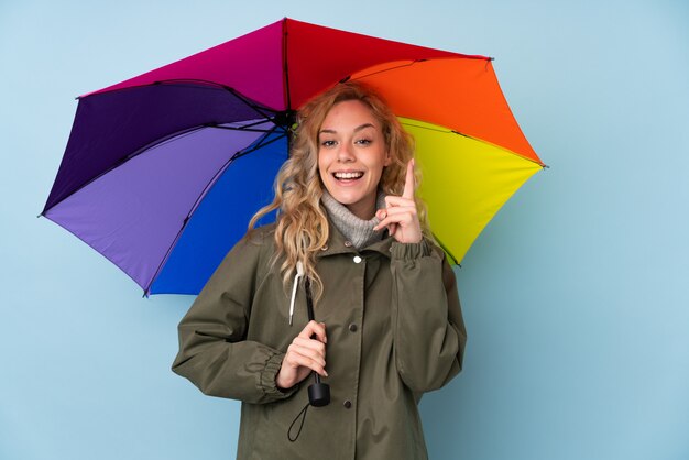 Junge blonde Frau, die einen Regenschirm lokalisiert auf blauer Wand hält, die eine große Idee zeigt