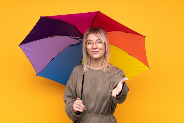 Junge blonde Frau, die einen Regenschirm über isoliertem gelbem Wandhandschlag nach vielem hält