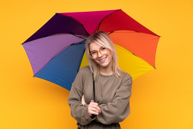 Junge blonde Frau, die einen Regenschirm über isoliertem gelbem Wandlachen hält