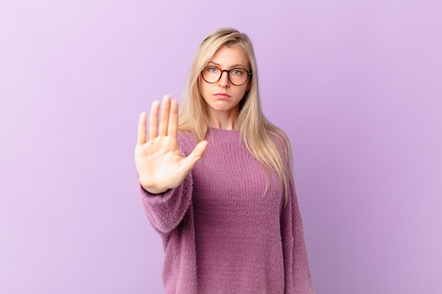 Junge blonde Frau, die ernst aussieht und offene Handfläche zeigt, die Stopp-Geste macht