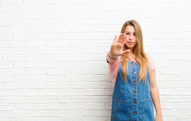 Junge blonde Frau, die ernst, streng, unzufrieden und wütend aussieht und offene Handfläche zeigt, die Stoppgeste über Ziegelmauer macht