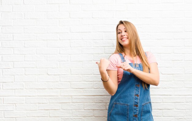 Junge blonde Frau, die fröhlich lächelt und beiläufig zeigt, um Raum auf der Seite zu kopieren, sich glücklich und zufrieden gegen Backsteinmauer fühlend