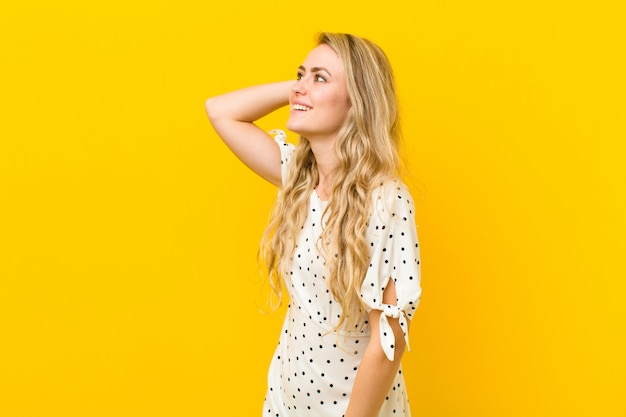 Junge blonde Frau, die fröhlich und beiläufig lächelt und Hand zum Kopf mit einem positiven, glücklichen und selbstbewussten Blick gegen gelbe Wand nimmt