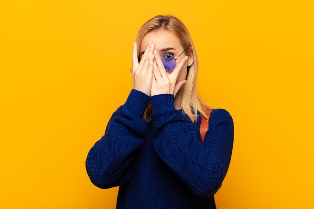 Junge blonde Frau, die Gesicht mit Händen bedeckt, zwischen Fingern mit überraschtem Ausdruck späht und zur Seite schaut