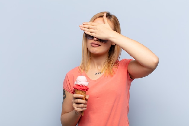 Junge blonde Frau, die Gesicht mit Hand bedeckt und ein Eis hält