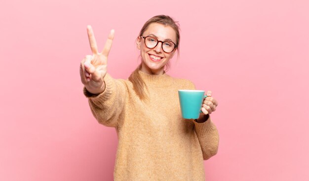 Junge blonde Frau, die glücklich, sorglos und positiv lächelt und aussieht und mit einer Hand Sieg oder Frieden gestikuliert. Kaffeekonzept