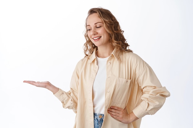 Junge blonde Frau, die ihre Handfläche mit Produkt betrachtet, Artikelexemplar zeigt, Produkt in der Hand ausstellt und zufrieden lächelt, über weißer Wand stehend