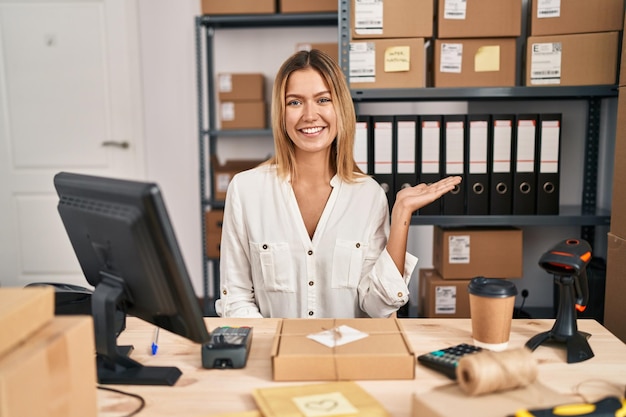 Junge blonde Frau, die im E-Commerce für kleine Unternehmen arbeitet, lächelt fröhlich, präsentiert und zeigt mit der Handfläche in die Kamera.