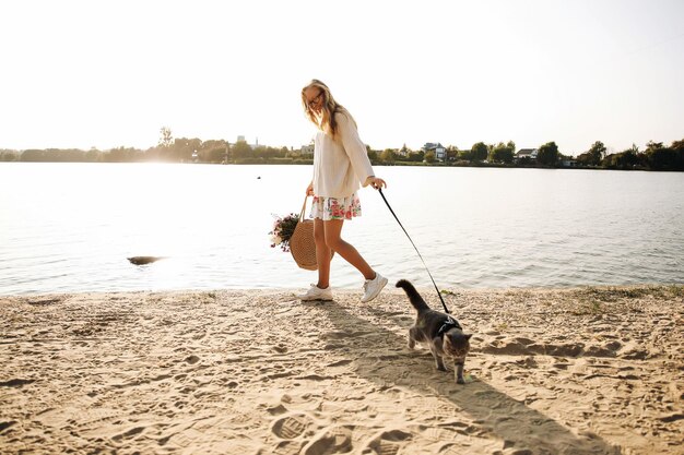 Junge blonde Frau, die im Frühling in einem Kleid und einem weißen Pullover mit einer schottischen geraden Katze an einem sonnigen Tag am Strand spazieren geht