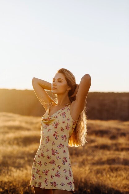 Junge blonde Frau, die in einem Feld mit trockenem Gras im Sonnenuntergangslicht steht