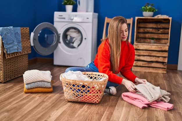 Junge blonde Frau, die Kleidung faltet und im Waschzimmer auf dem Boden sitzt