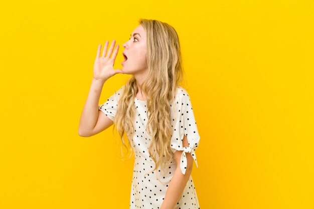 Junge blonde Frau, die laut und wütend schreit, um Platz auf der Seite zu kopieren, mit Hand neben Mund gegen gelbe Wand