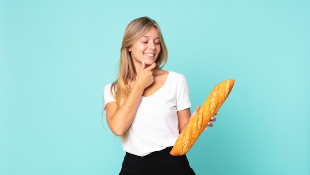 Junge blonde Frau, die mit einem glücklichen, selbstbewussten Ausdruck mit der Hand am Kinn lächelt und ein Brotbaguette hält