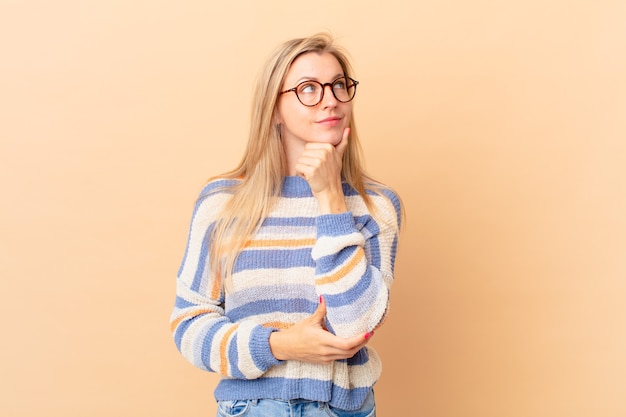 Junge blonde Frau, die mit einem glücklichen, selbstbewussten Ausdruck mit der Hand am Kinn lächelt