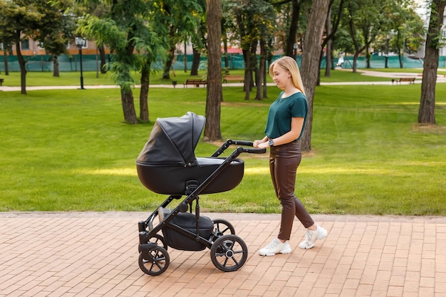 Junge blonde Frau, die mit schwarzem Kinderwagen im Sommerpark spaziert, glückliche Mutter mit Baby im Kinderwagen?