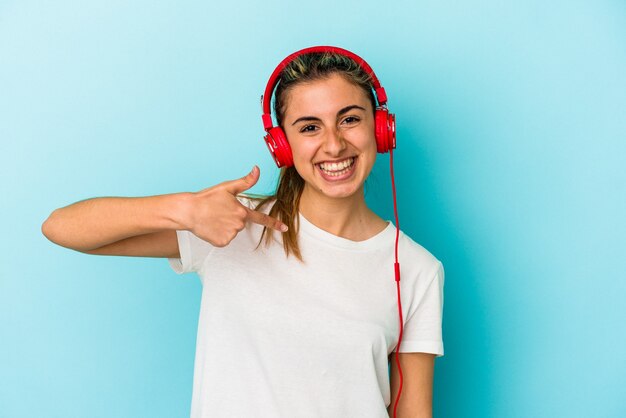 Junge blonde Frau, die Musik auf Kopfhörern lokalisiert auf blauem Hintergrund Person hört, zeigt von Hand auf einen Hemdkopierraum, stolz und zuversichtlich